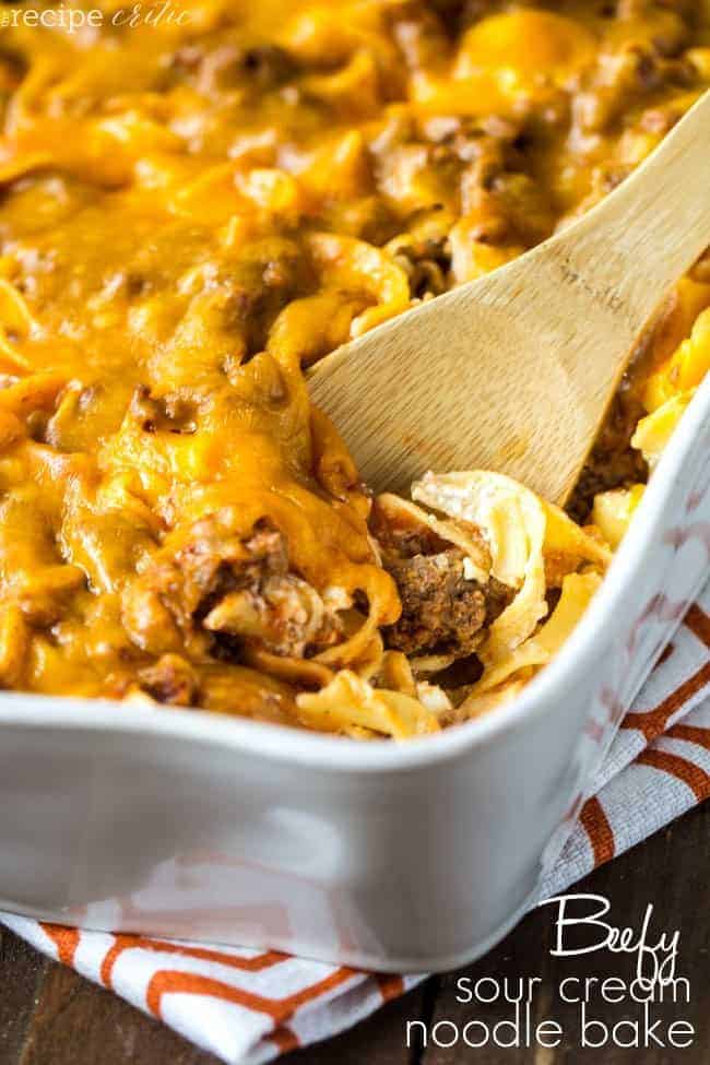 Delicious and cheesy casserole with a sour cream mixture that the entire family will love! Beefy Sour Cream Noodle Bake in a white casserole dish with a wood spoon.