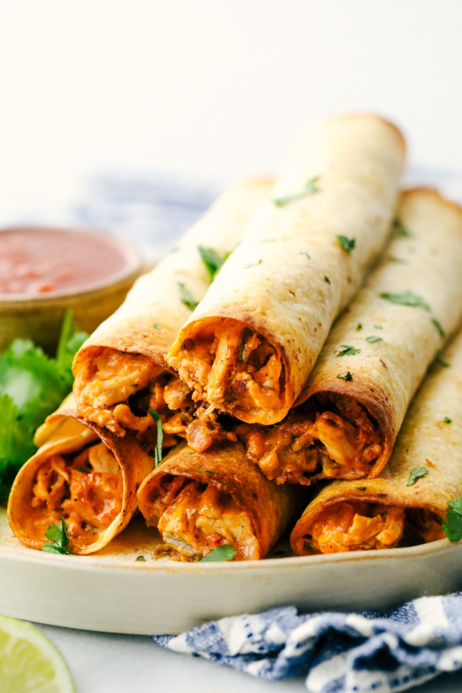 Cream cheese chicken taquitos are filled with a tender chicken mixture with cheese sprinkled over top then rolled in a flour tortilla and baked to perfection. These taquitos are a family favorite! Chicken taquitos stacked on top of each other on a plate.