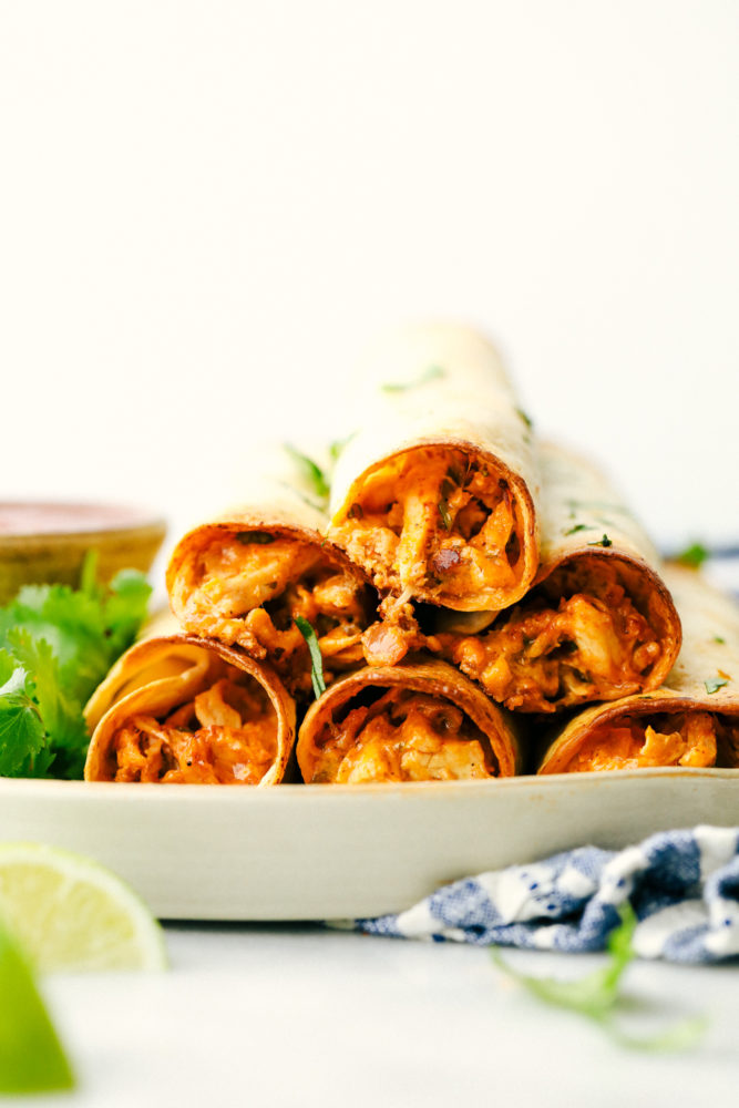 Cream cheese chicken taquitos are filled with a tender chicken mixture with cheese sprinkled over top then rolled in a flour tortilla and baked to perfection. These taquitos are a family favorite! Cream cheese chicken taquitos are stacked on top of each other with lime wedges as a garnish.