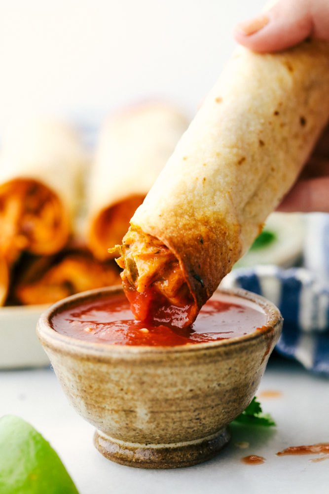 Cream cheese chicken taquitos are filled with a tender chicken mixture with cheese sprinkled over top then rolled in a flour tortilla and baked to perfection. These taquitos are a family favorite! Cream cheese chicken taquito is being dipped in a red salsa.