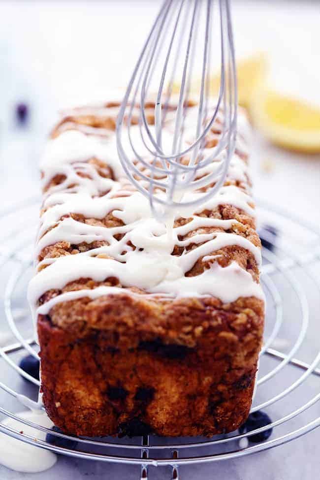 An amazingly moist muffin bread filled with blueberry pie filling. Topped with a crumb topping and drizzled in a lemon glaze this bread is incredible! Blueberry pie bread on a cooling rack with lemon glaze being spread by a whisk.