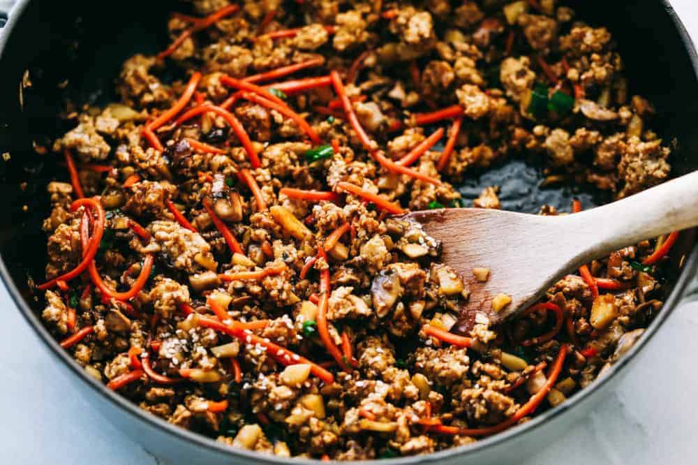 Asian turkey lettuce wraps are packed with intense flavor from tender meat and seasoned, crunchy veggies. These light and tasty wraps are the perfect healthy appetizer or even dinner! The filling for asian turkey lettuce wraps in a skillet with a wooden spoon.