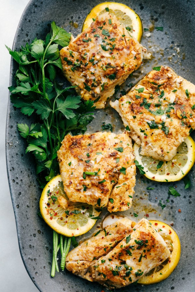 Baked Cajun Garlic Butter Cod is a tender and juicy cod filet baked in a buttery cajun seasoning with garlic cloves and olive oil making this cod a flavorful and delicious dinner. Baked to perfection in 15 minutes! Baked cajun garlic butter cod served on a black speckled plate with green garnish and lemon slices underneath the cod filets.