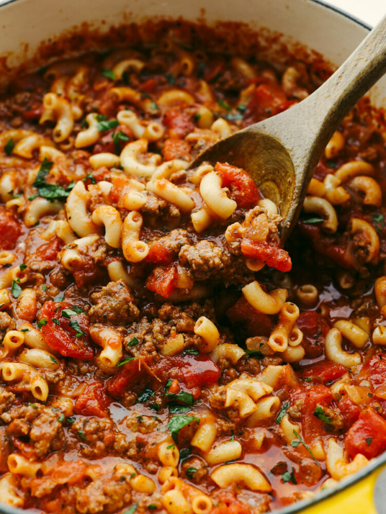 American Goulash is an easy old-fashioned comforting meal that is perfect for the cold winter months. Everything cooks in the same pot, even the pasta! A pot with American goulash being stirred with a wooden spoon.